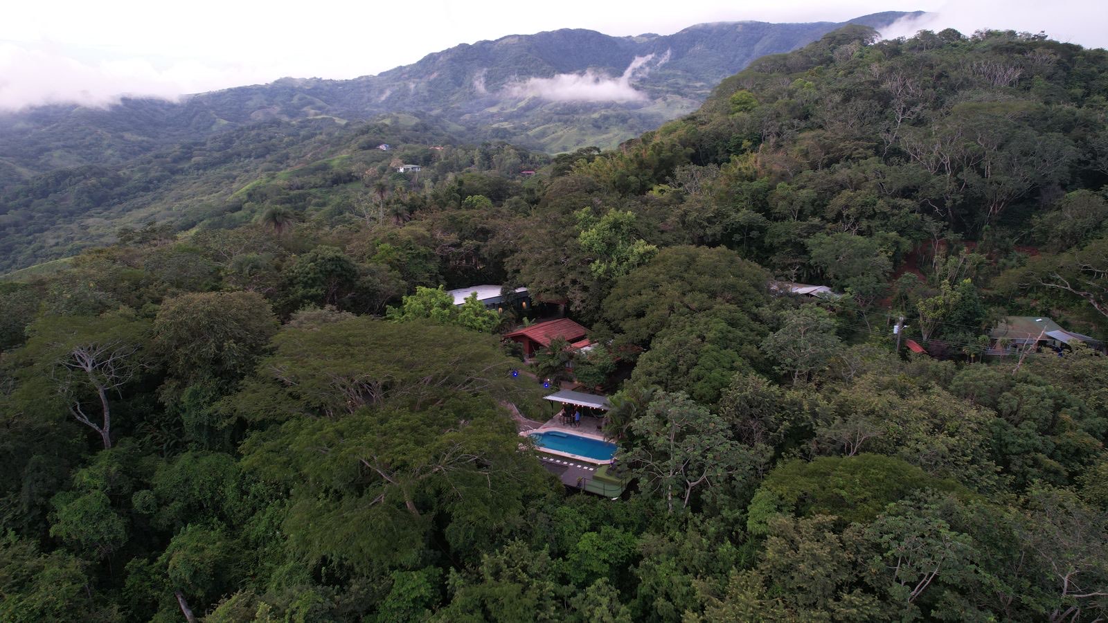 Vista de dron: el sitio es un claro dentro del bosque, con la piscina y los techos entre los árboles y las montañas de Montes de Oro detrás